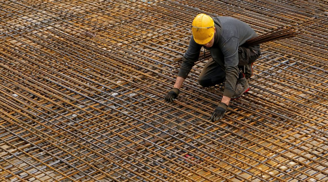 Bürokratie Baubranche, hier: Bauarbeiter mit gelbem Schutzhelm kniet auf großflächigen Armierungsgittern